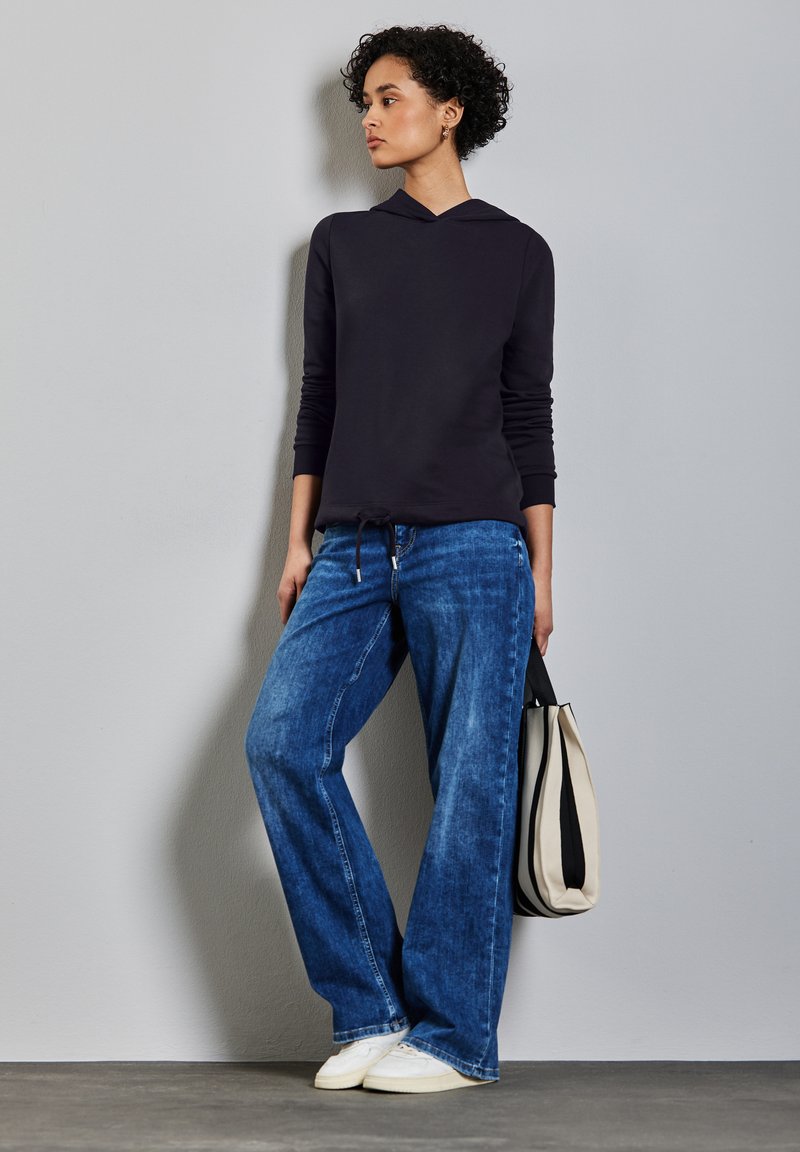 Schwarzer Kapuzenpullover mit langen Ärmeln, kombiniert mit weit geschnittenen blauen Jeans. Hält eine gestreifte Tasche in Schwarz und Creme.