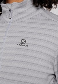 Světlé šedé zipové sako z měkké tkaniny. Má texturovaný pruhovaný vzor a malé černé logo Salomon na hrudi.