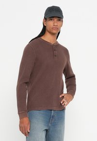 Mann mit langen Zöpfen, der eine schwarze Kappe, ein braunes Waffelstrick-Hemdkleid mit langen Ärmeln und ausgebleichte blaue Jeans trägt, steht vor einem weißen Hintergrund.