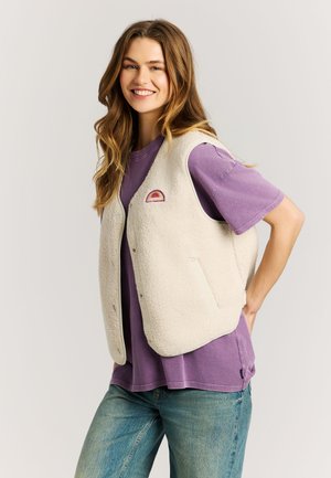 Glunderende vrouw met lang haar draagt een paarse t-shirt, een wit sherpa-vest met een klein zonembleem en lichtblauwe jeans, staand met de hand op de heup.