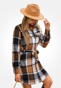 Abito a quadri marrone, nero e bianco con maniche lunghe, chiusura frontale a bottoni e cintura. Abbinato a un cappello a tesa larga color beige. Motivo a quadri notevole.