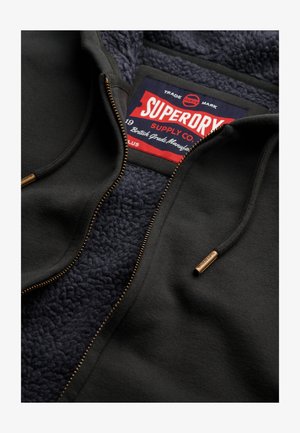 Schwarze Zip-Jacke mit marineblauer Fleece-Innenfütterung, markiertem Etikett, goldenem Reißverschluss und Kordelzug-Akzenten. Besteht aus weichem, strukturiertem Stoff.