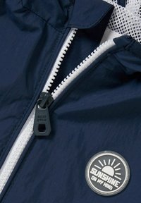 Lekka granatowa kurtka z białym suwakiem, plastikową naszywką z logo w kształcie słońca oraz detalem podszewki z siateczki. Pokazano teksturowaną tkaninę.