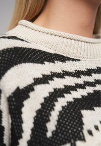 Stickad tröja med ett svart och vitt zick-zack-mönster, ribbad rund halsringning och textilier med struktur. Närbild av designen.