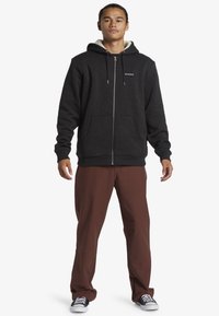 Felpa nera con zip, tasche laterali e cappuccio con cordino, caratterizzata da un morbido rivestimento interno, abbinata a pantaloni marroni e scarpe nere.
