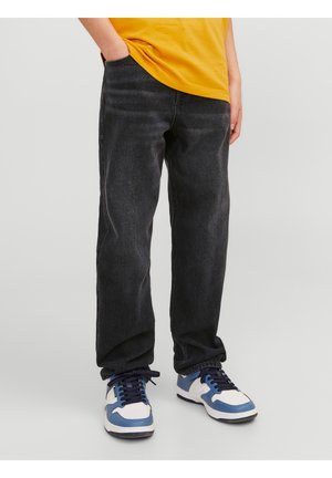Person iført løsthængende sorte jeans, gul skjorte og hvide sneakers med blå og sorte detaljer, stående med den ene hånd i lommen.