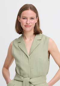 Tuta senza maniche, con bottoni, in tessuto verde chiaro, dotata di vita cinturata, colletto e superficie liscia e strutturata.