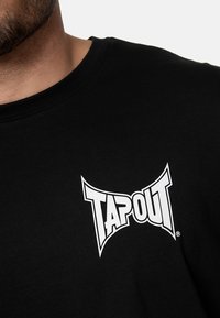 Fekete pamut póló, amelyen a bal mellen egy kiemelkedő fehér "TAPOUT" logó található. Kerek nyakkivágású, rövid ujjú fazon.