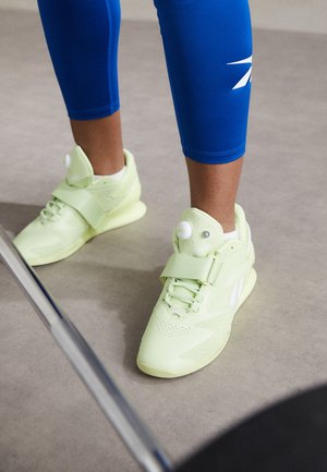 Personne portant des baskets Reebok jaune pâle et des leggings bleus debout sur un sol gris à côté d'une patte de chaise en métal.