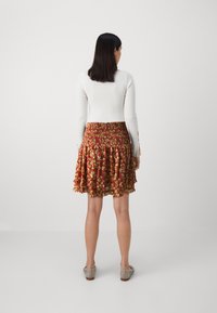 Jupe florale rouge avec un ourlet volanté, dotée d'une taille froncée. Un haut à manches longues blanc est porté avec elle. Des chaussures de couleur neutre sont visibles.