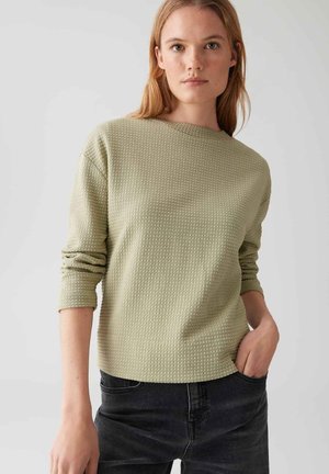 Woman with light brown hair wearing a textured sage green long-sleeve top and dark jeans, hand in pocket, against a plain light background.