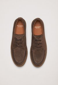 Chaussures en suède marron à lacets avec un bout arrondi, présentant des accents cousus et une semelle intérieure en cuir beige. Texte de la marque visible à l'intérieur.