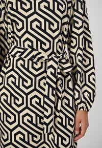 Schwarzes und beiges geometrisch gemustertes Kleid mit langen Ärmeln und einem gebundenen Taillengürtel, teilweise wird eine rechte Hand an der Seite gezeigt.