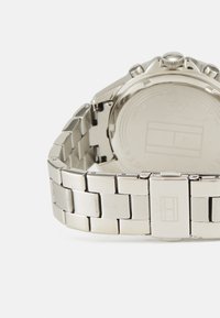 Montre en acier inoxydable avec une finition brossée et polie. Elle présente un bracelet à maillons et une fermeture sécurisée. Le dos affiche des détails en relief.