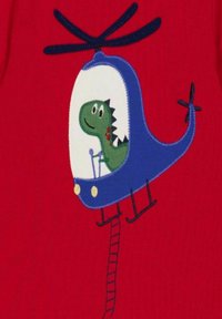 Modrý vyšitý vrtulník s pilotem zeleného dinosaura. Pozadí je červené, obsahuje detail žebříku a design usmívajícího se dinosaura.