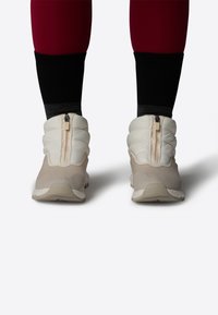 Lyse beige polstrede sneakers med lynlås, tekstureret overdel og fleksible såler. Båret med sorte og grå sokker, hvilket viser et sporty design.