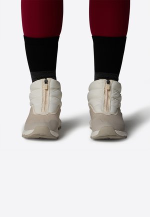 Lyse beige polstrede sneakers med lynlås, tekstureret overdel og fleksible såler. Båret med sorte og grå sokker, hvilket viser et sporty design.