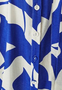 Mėlynas marškinėlių mygtukais, papuoštas baltais abstrakčiais raštais, su perlų blizgesio mygtukais ir tekstūruotu audiniu.