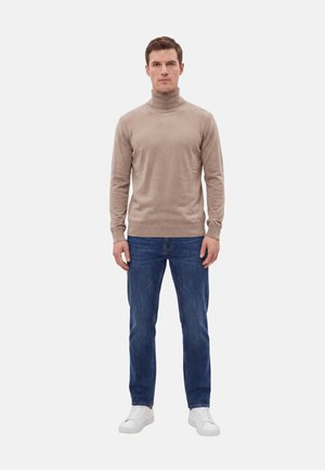 Uomo in piedi rivolto in avanti, indossa un maglione a collo alto beige, jeans blu e sneakers bianche su sfondo semplice.