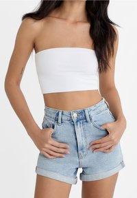 Mujer que lleva un top corto sin mangas blanco y pantalones cortos de mezclilla azul claro, enrollados, con las manos en los bolsillos, frente a un fondo liso.