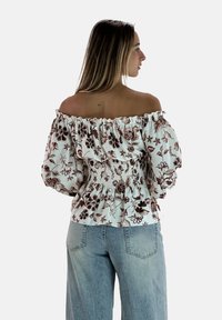 Blusa bianca off-the-shoulder con stampa floreale marrone, caratterizzata da un corpetto arricciato e maniche a palloncino, abbinata a jeans blu chiaro.
