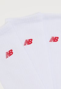 Τρεις λευκές κάλτσες με κόκκινα λογότυπα New Balance κεντημένα κοντά στον αστράγαλο, τοποθετημένες η μία δίπλα στην άλλη σε λευκό φόντο.