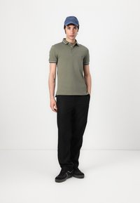 Ελιά πράσινο πόλο μπλουζάκι με αντίθετες σκούρες ρίγες στον γιακά, σε συνδυασμό με μαύρο παντελόνι και μαύρα αθλητικά παπούτσια, ολοκληρωμένο με μπλε καπέλο.