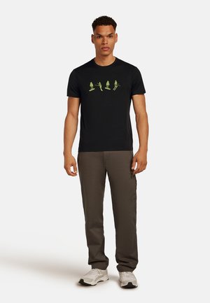 Man in een zwart t-shirt met groene skifiguren, bruine broek en witte sneakers, staand tegen een effen witte achtergrond.