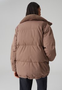 Doudoune marron avec un col en fourrure douce. Design matelassé, coupe surdimensionnée et poignets côtelés. Vue de dos montrant une texture lisse et un ourlet arrondi.