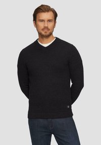 Schwarzer Strickpullover mit V-Ausschnitt, texturierter Oberfläche und geripptem Saum. Über einem weißen Hemd getragen, kombiniert mit dunklen Jeans.