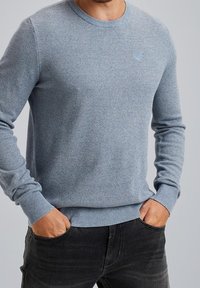 Pull tricoté bleu clair avec col rond, motif texturé, manches ajustées et petit logo sur la poitrine ; associé à un pantalon noir.