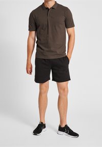 Ciemnobrązowa koszulka polo z kołnierzykiem i zapięciem na dwa guziki, zestawiona z czarnymi szortami. Model nosi czarne sportowe buty.