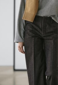 Pantalon marron foncé à la coupe structurée avec des plis marqués, associé à un haut à manches longues gris clair avec une manche côtelée beige.