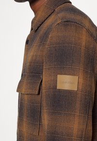 Brun och marinblå rutig flanellskjorta med krage, knappstängning och en märklogotyp på bröstfickan. Texturerad tyg med lätt glans.