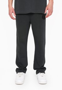 Dark gray athletic pants with a fitted design, featuring a textured fabric, side stripes, and a logo patch on the left thigh. Worn with white sneakers.