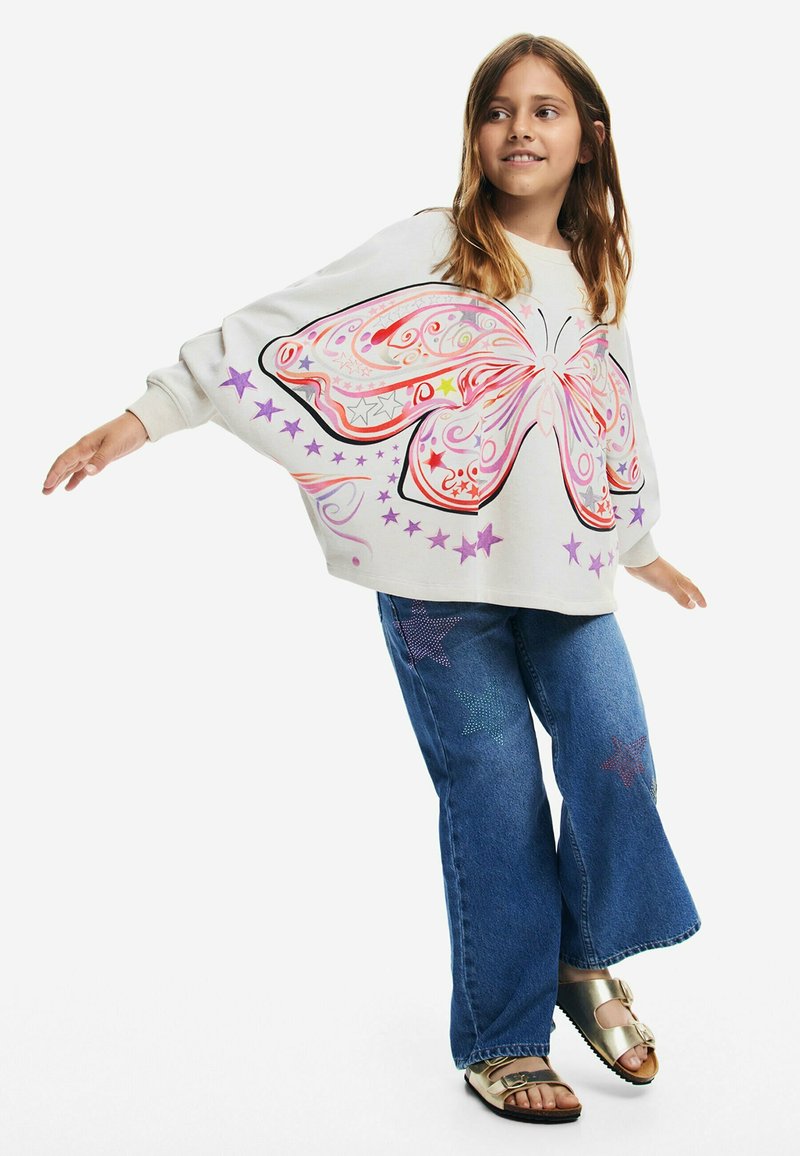 Felpa grigia chiara con una grande grafica di una farfalla multicolore, abbinata a jeans a campana blu con decorazioni a forma di stella e sandali dorati.