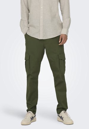 Hombre con pantalones cargo verde oliva, camisa beige abotonada y zapatillas blancas con detalles en azul oscuro, de pie frente a un fondo liso.