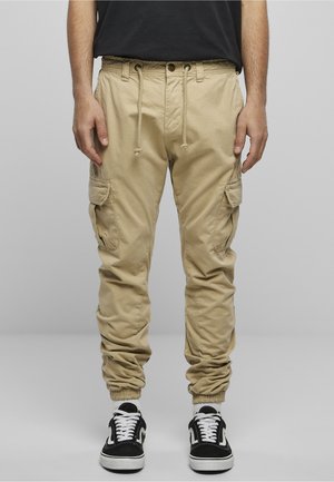 Un modèle court porte un jogger cargo beige avec cordon, poches latérales et poignets élastiques, associé à une chemise noire et des baskets noir et blanc.