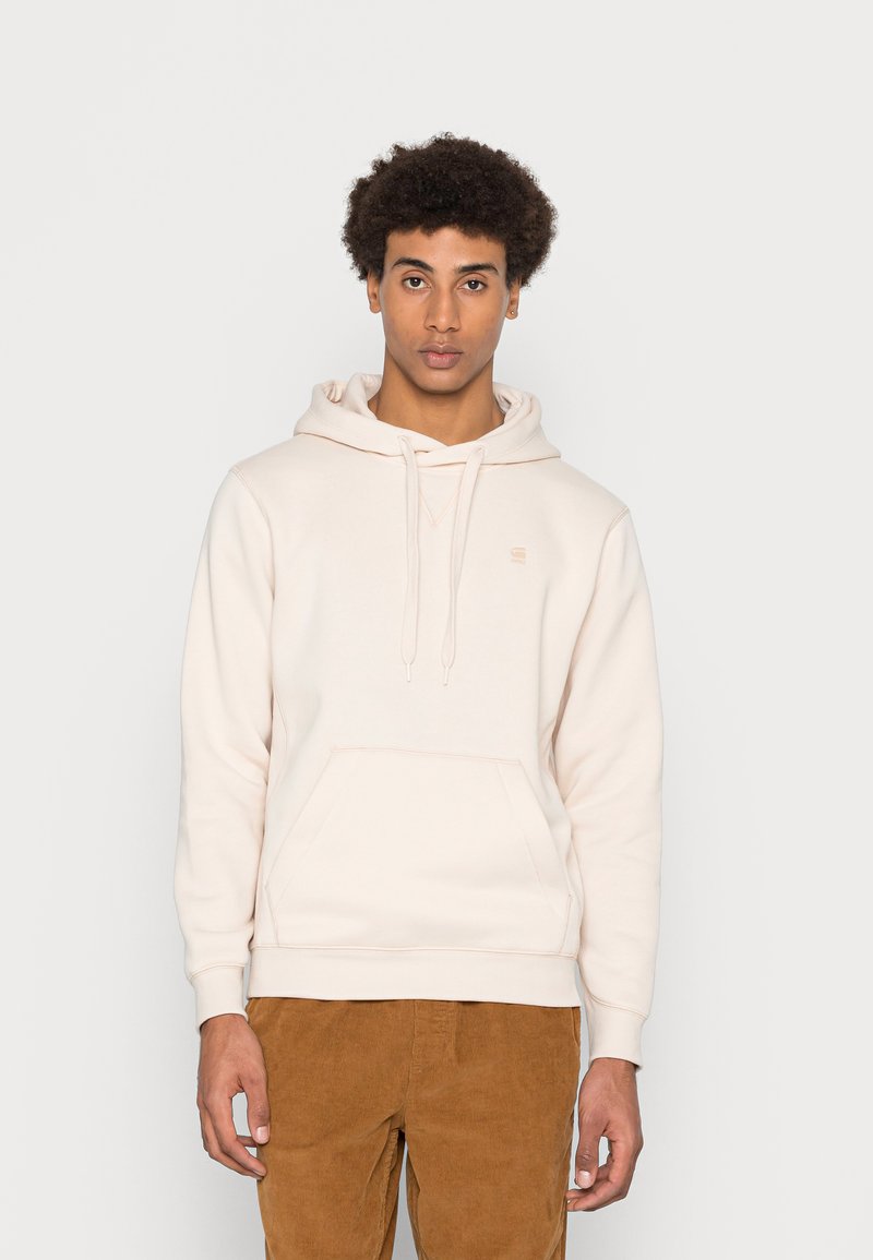 Fleece em tons de bege, feito de um tecido macio, com um bolso canguru, capuz com cordão e um logotipo minimalista no peito. Combinado com calças de sarja castanhas.