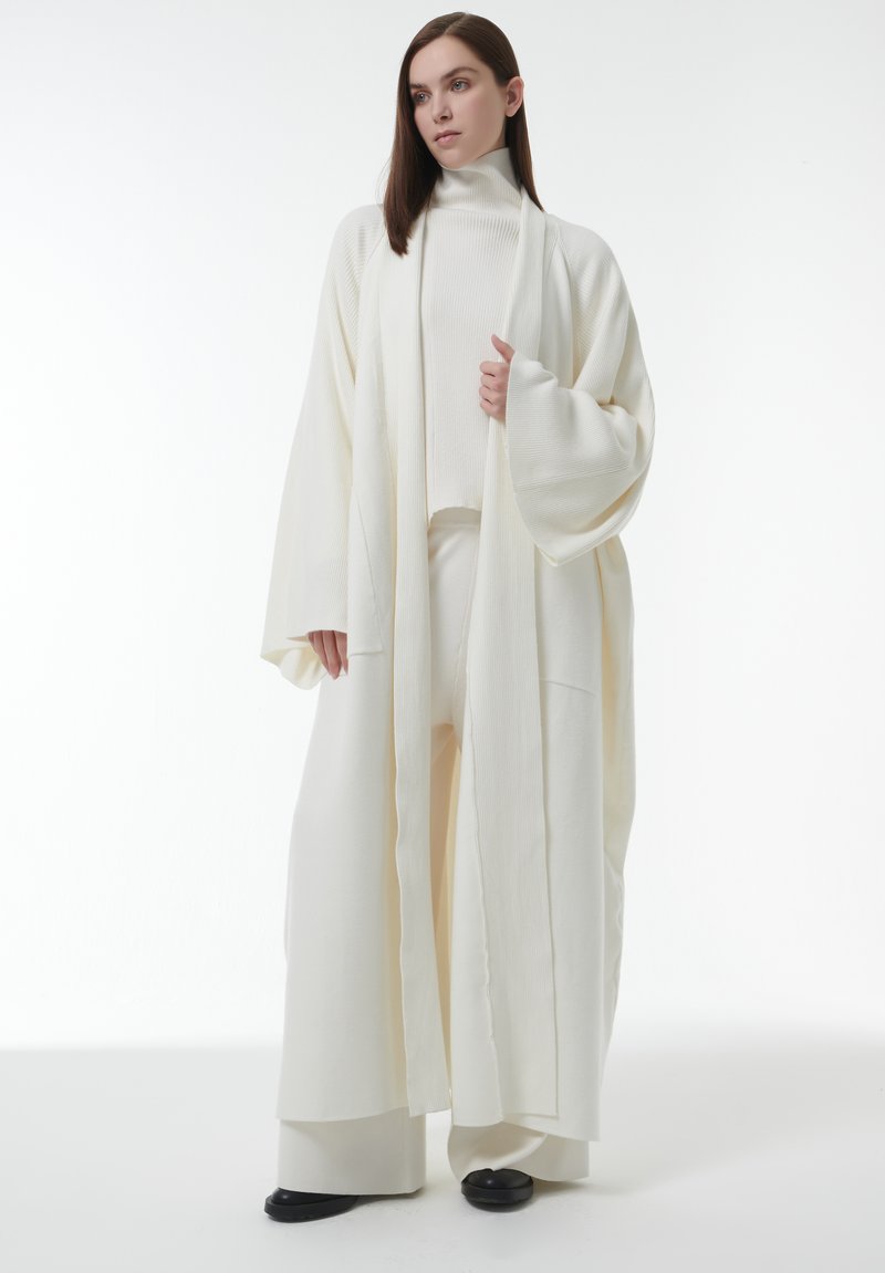 Lange roomkleurige robe met brede mouwen, met een zachte textuur en een open voorkant. Ggedragen over een bijpassend aansluitend top en wijde broek.