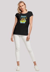 Svart bomull T-skjorte med et fargerikt "Scooby-Doo!" motiv. Designet inkluderer blomster og et lyst gult element. Kombinert med hvite bukser.