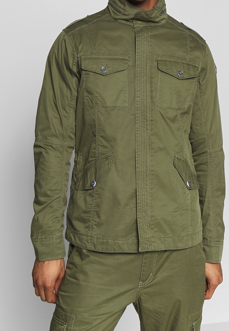 Veste vert olive avec un col haut, dotée de deux poches sur la poitrine et de deux poches latérales, fabriquée en tissu léger avec des coutures visibles.