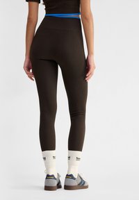 Bruine geribde leggings met een hoge tailleband, voorzien van een blauwe acccent. Het model draagt witte sokken en grijs-blauwe sneakers.