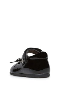 Zapato de charol negro con punta redondeada, tira de Velcro y detalle de lazo en la parte frontal. Textura suave y tacón bajo.