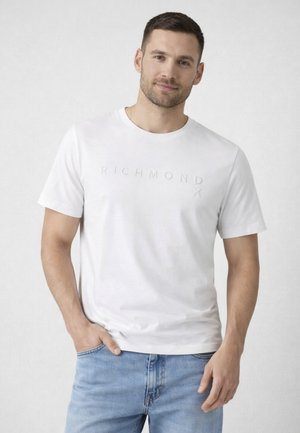 Uomo che indossa una t-shirt bianca Richmond X e jeans azzurri, in piedi con una mano nella tasca davanti a uno sfondo chiaro e uniforme.