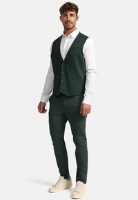 Grønn teksturert vest med fem knapper, kombinert med matchende bukser og hvit skjorte. Modellen har på seg lyse sneakers.