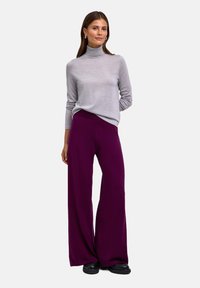 Maglione grigio a collo alto abbinato a pantaloni viola a vita alta e gamba larga, caratterizzati da una trama liscia e una vestibilità rilassata. Calzature nere completano il look.
