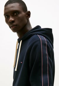 Marineblauwe hoodie van katoen, met een crèmekleurige trekkoord. Voorzien van zijstrepen in rood en wit, met een klein logo op de borst.