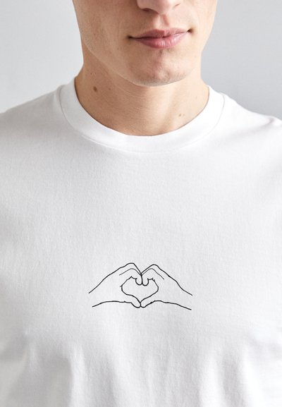 Camiseta de algodón blanca con un dibujo en línea negra de dos manos formando un corazón en la zona del pecho. Diseño clásico de cuello redondo.