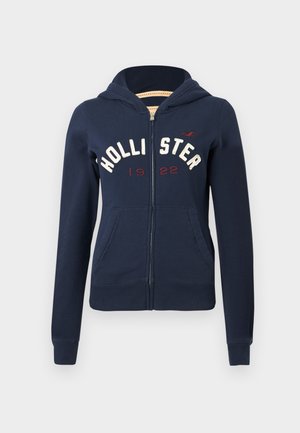 Tummansininen vetoketjullinen huppari etutaskuilla, valkoinen "HOLLISTER" -teksti, punaiset "1922"-numerot ja huppu yksinkertaisella taustalla.
